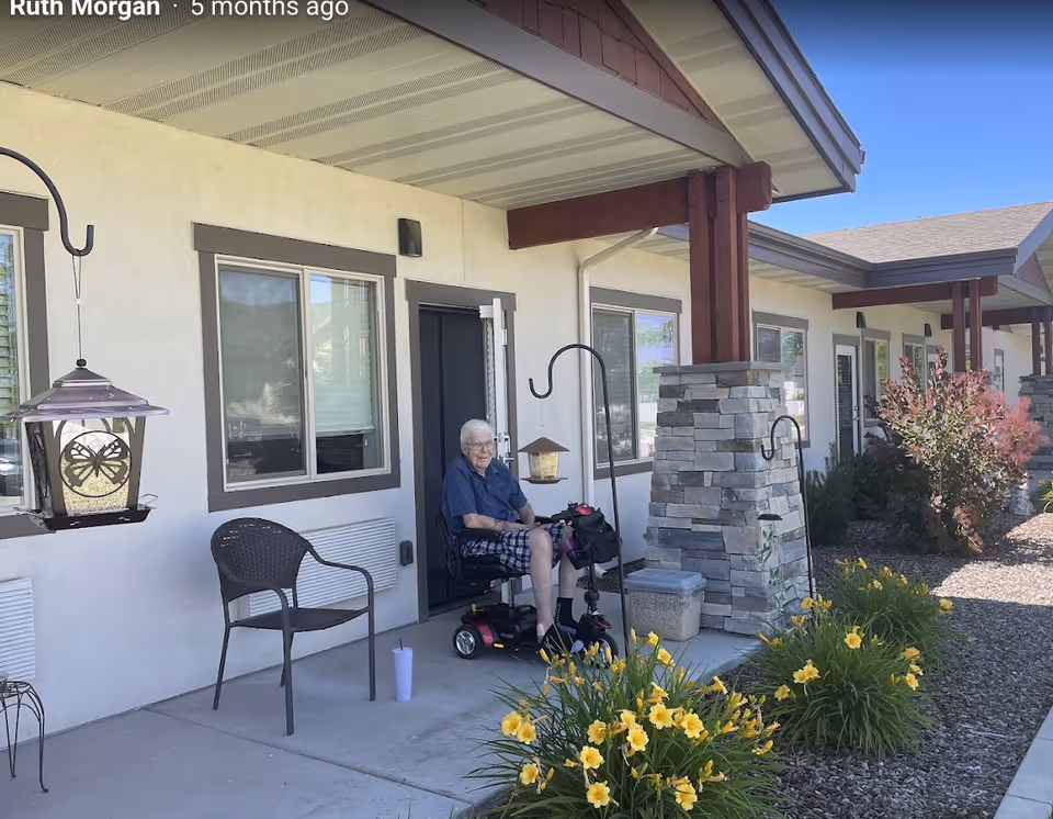 An elderly man sitting on a mobility scooter on the covered patio of a senior living facility. The patio has a chair, a cup on the ground, hanging bird feeders, and is decorated with yellow flowers and shrubs. The building exterior is light-colored with brown trim and stone pillars supporting the roof.