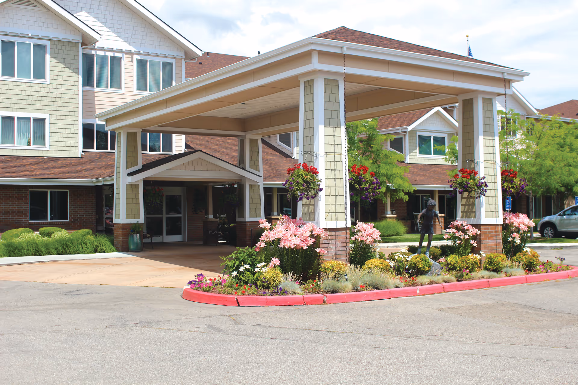 Entrance of a senior living facility with a covered drop-off area supported by four pillars. The area is surrounded by well-maintained flower beds with pink and red flowers, hanging flower baskets, and greenery. The building has multiple windows and a mix of brick and siding exterior.