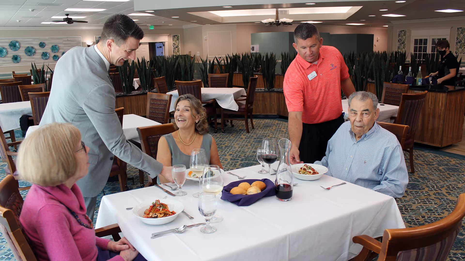 Two elderly individuals seated at a dining table with plates of food and drinks, being served by two staff members in a well-lit dining room with tables, chairs, and decorative plants in the background.