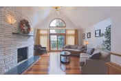 A spacious living room with a high vaulted ceiling, large arched window with curtains, a stone fireplace decorated with a wreath, hardwood floors, two gray sofas, a round coffee table, a black armchair, framed pictures on the wall, and a potted plant in the corner.