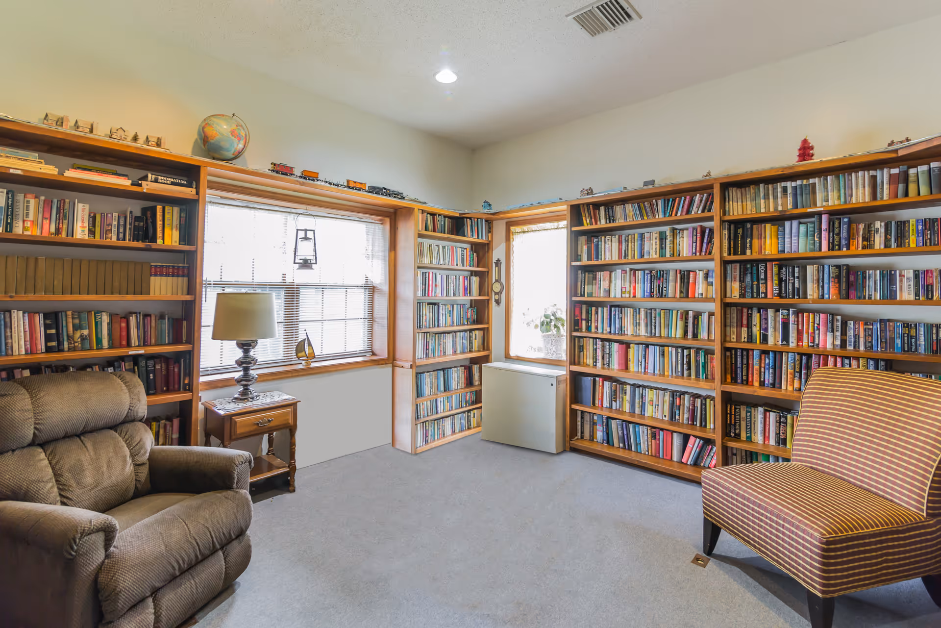 A cozy reading room with carpeted floor, featuring multiple wooden bookshelves filled with books. There is a large window with blinds letting in natural light, a small wooden side table with a lamp and decorative items, a comfortable recliner chair, and a striped upholstered chair.