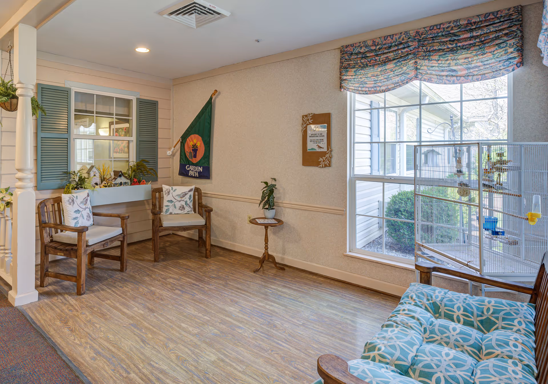 A cozy seating area in a senior living facility with wooden chairs featuring floral cushions, a small round table with a potted plant, a large window with patterned valance, and a birdcage. The walls are decorated with a flag and a bulletin board, and there is a decorative window frame with shutters and plants on the left side.