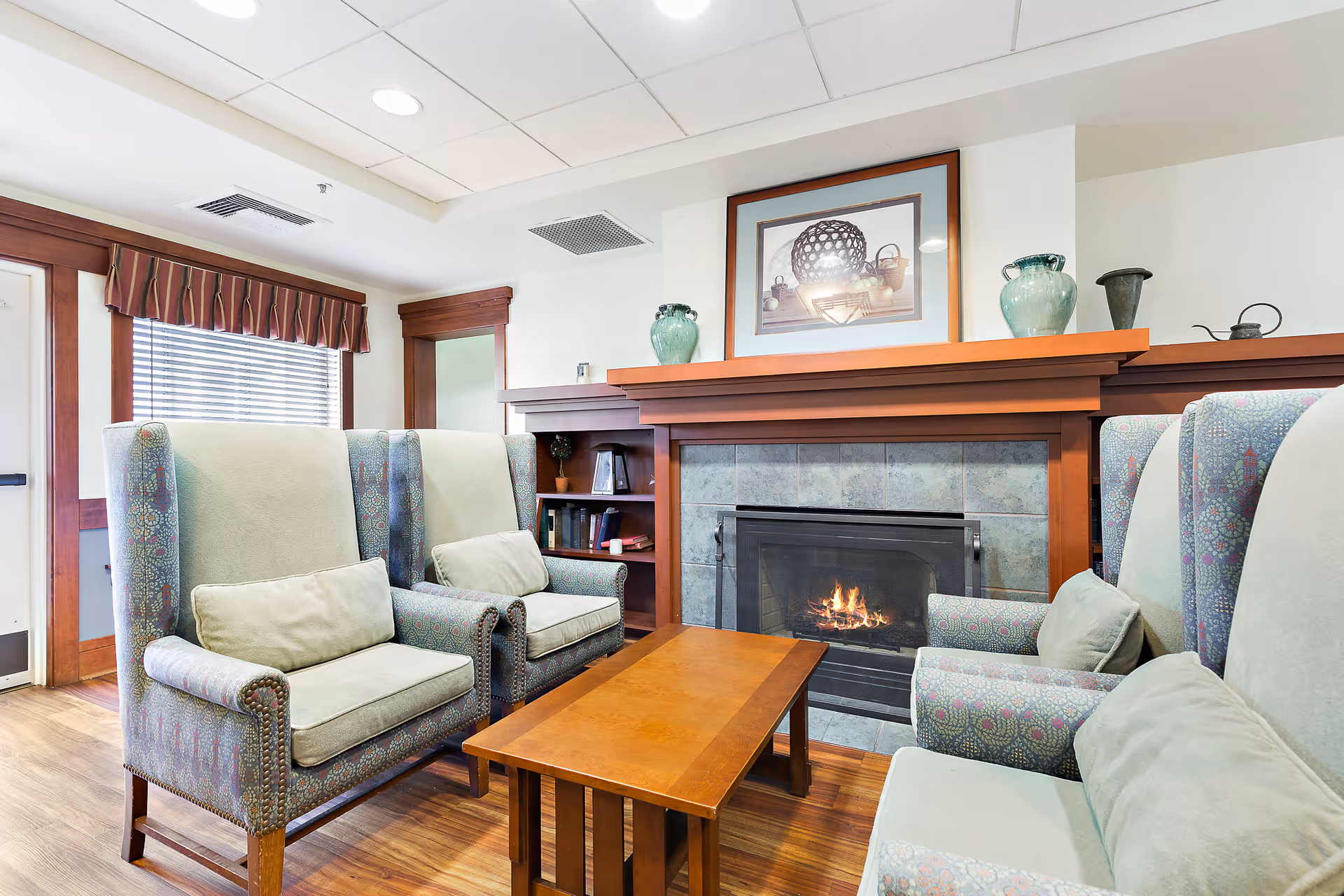 A cozy living room area with four upholstered armchairs arranged around a wooden coffee table in front of a lit fireplace. The fireplace has a wooden mantle decorated with vases and a framed picture above it. The room has wooden flooring, a window with blinds and a valance, and built-in shelves with books and decorative items.