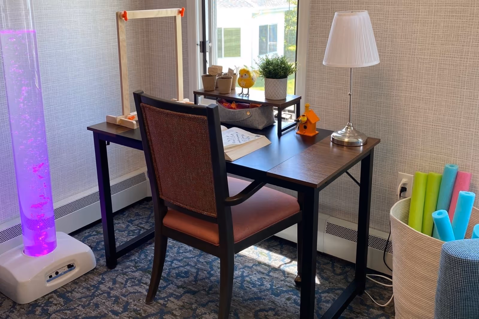 Small activity desk with a chair, lamp, craft supplies and a purple bubble tube by a window in a senior living facility room.