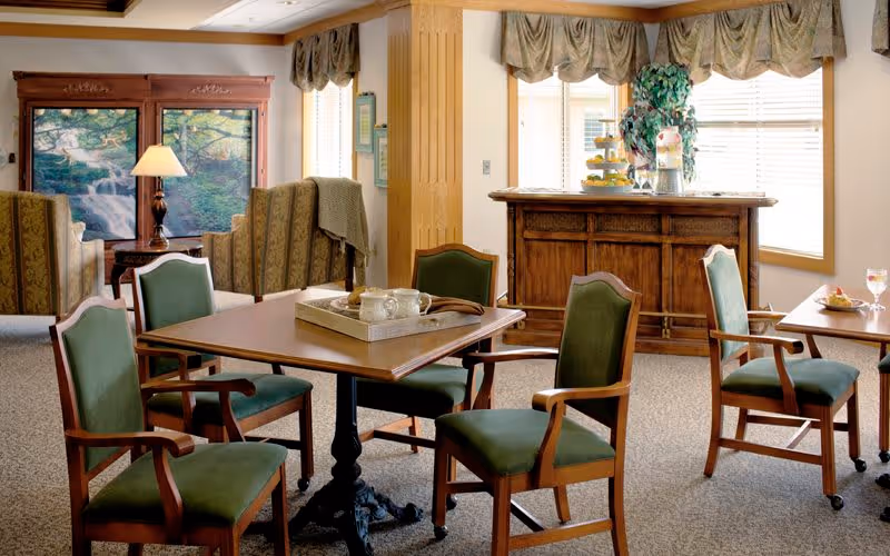 A cozy common area with wooden furniture including a table surrounded by four green cushioned chairs. In the background, there are two armchairs facing a large window with a scenic waterfall view. A wooden cabinet with decorative items and a tiered tray is positioned near windows with valance curtains, allowing natural light to fill the room.