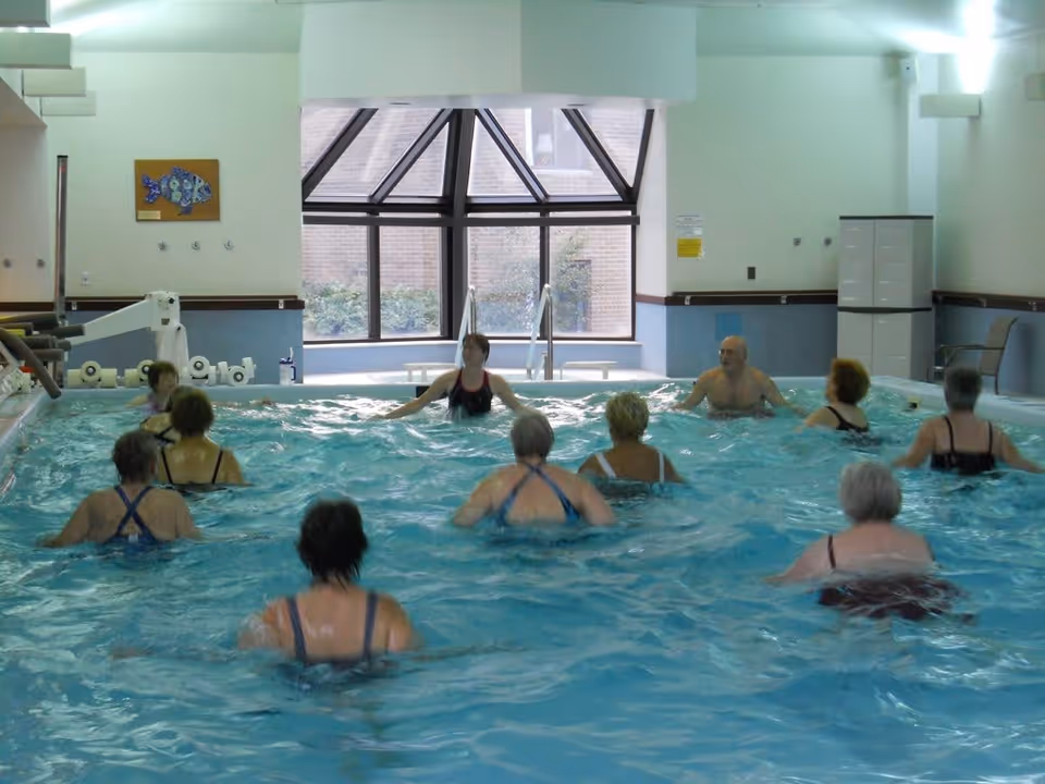 A group of elderly people participating in a water exercise class in an indoor swimming pool with large windows and a glass ceiling section in the background.