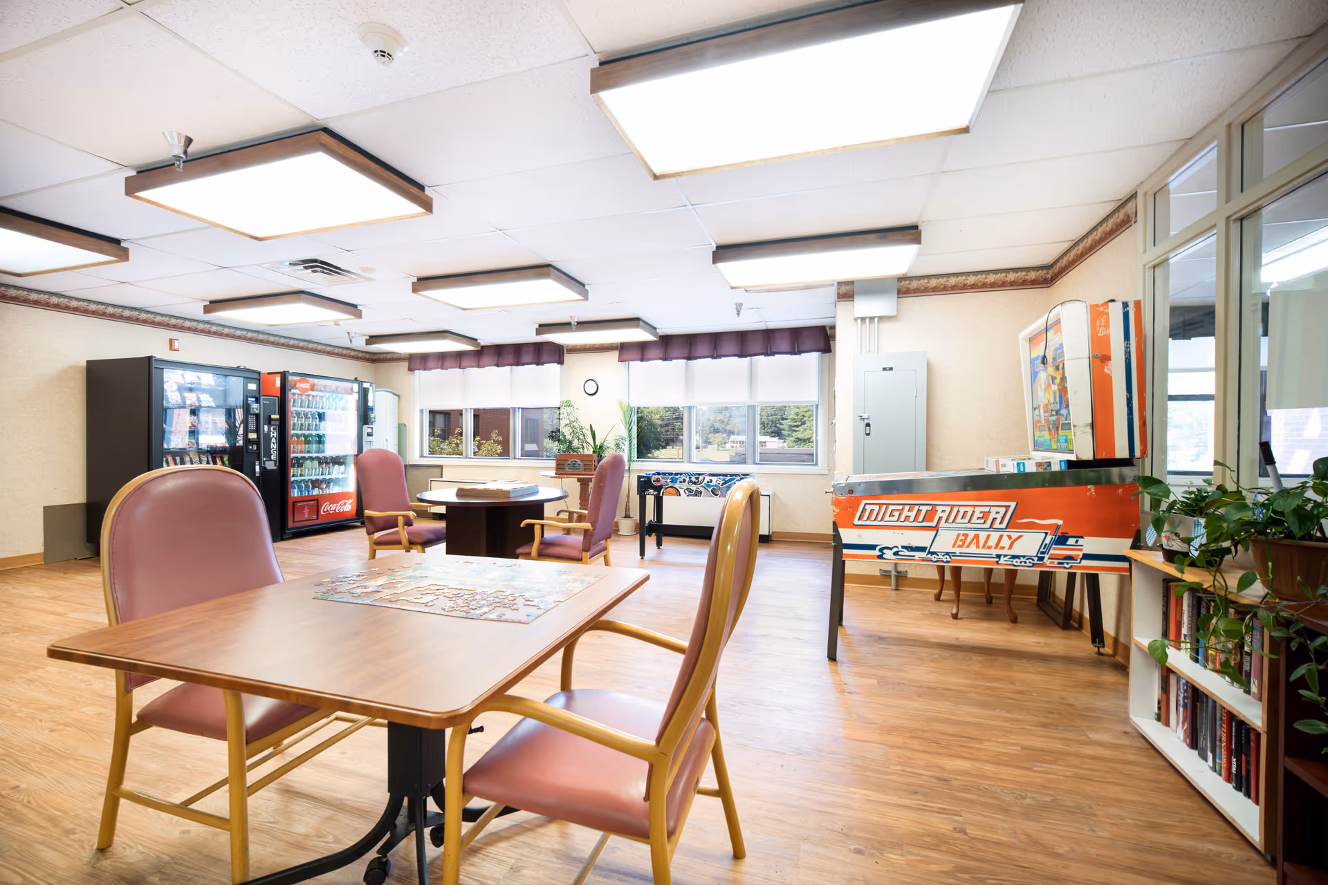A bright and spacious common room with wooden floors and ceiling lights. The room features several tables and chairs with pink cushions, a vending machine area with snacks and drinks, a foosball table, and a vintage pinball machine labeled 'Night Rider Rally'. There are large windows with purple valances letting in natural light, and some potted plants and bookshelves along the walls.