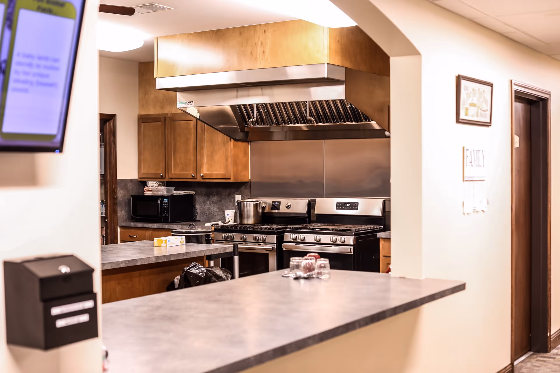 Interior view of a kitchen area with two stainless steel gas stoves, a large exhaust hood, wooden cabinets, a microwave, and a countertop with some glasses and a box on it. The kitchen is visible through an arched opening in the wall.