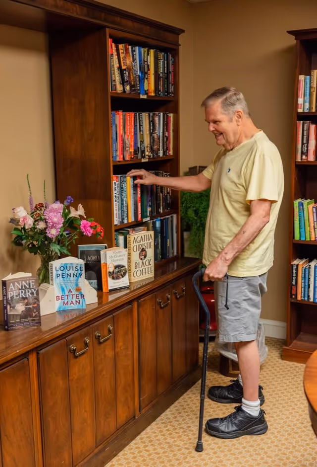 An elderly man with a cane is standing and smiling while selecting a book from a wooden bookshelf filled with various books. There are additional books and a vase with flowers displayed on the cabinet below the bookshelf. The room has a warm, cozy atmosphere with beige walls and carpeted flooring.