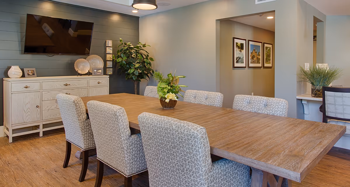 A dining room with a large wooden table surrounded by eight upholstered chairs with patterned fabric. A decorative plant centerpiece is on the table. In the background, there is a light-colored sideboard with decorative items and a flat-screen TV mounted on a dark accent wall. The room has wooden flooring and neutral-colored walls with framed pictures visible in an adjacent hallway.