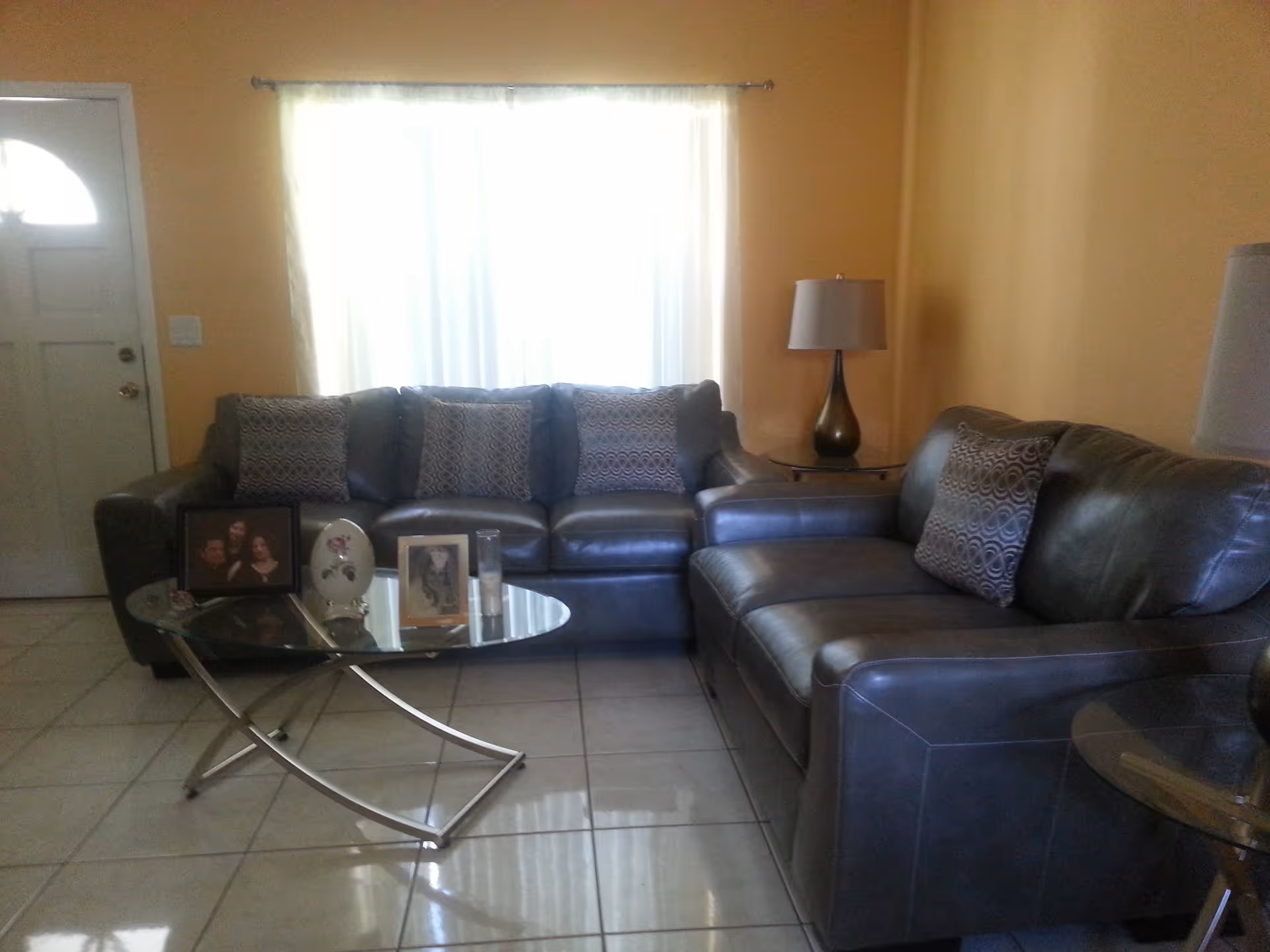 Sunlit living room with a gray leather sectional sofa, glass coffee table holding framed photos, and lamps by a curtained window and front door.
