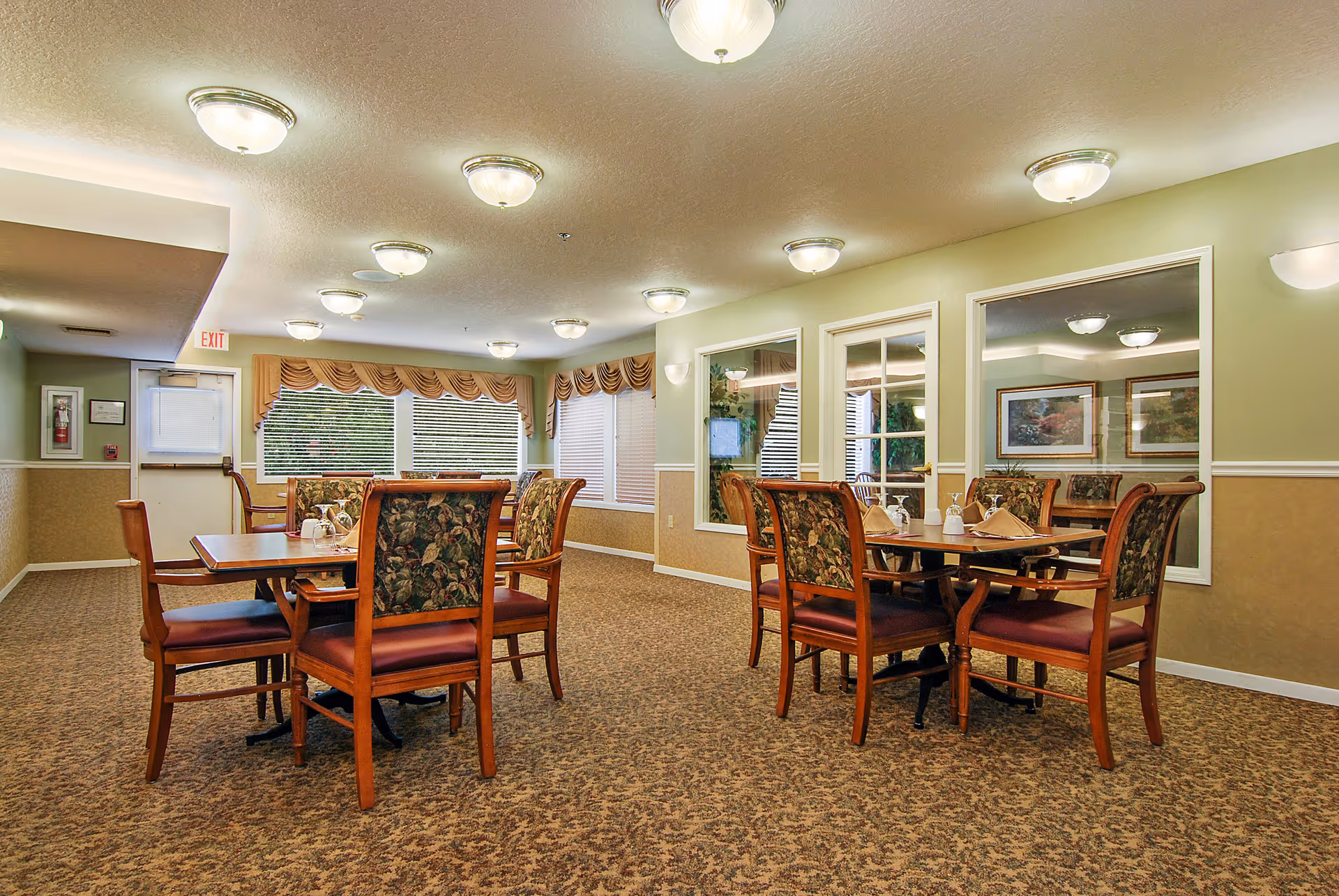 A well-lit dining room with wooden tables and upholstered chairs on patterned carpet beneath multiple ceiling lights and large windows.