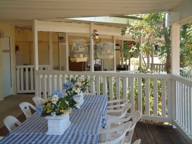 Covered outdoor patio area with a long table covered in a blue and white checkered tablecloth, decorated with flower arrangements. White plastic chairs surround the table. Bird cages and birdhouses hang from the ceiling. The patio is enclosed with white railings and overlooks greenery and trees.