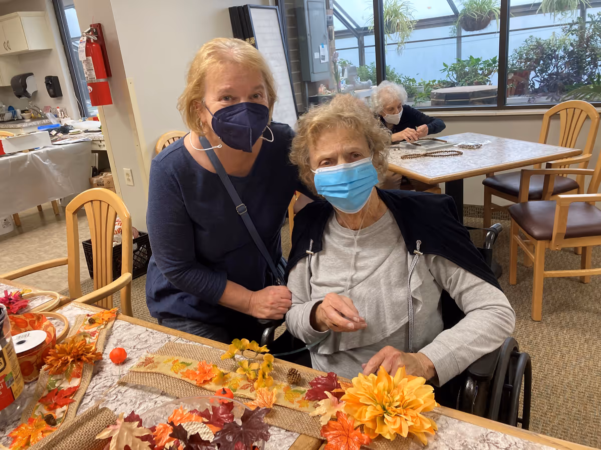 Two elderly women wearing face masks are seated at a table decorated with autumn-themed items including artificial leaves and flowers. One woman is sitting in a wheelchair while the other is leaning close to her. In the background, another elderly woman is seated at a separate table near large windows with plants visible outside. The room appears to be a communal indoor space with wooden chairs and tables.