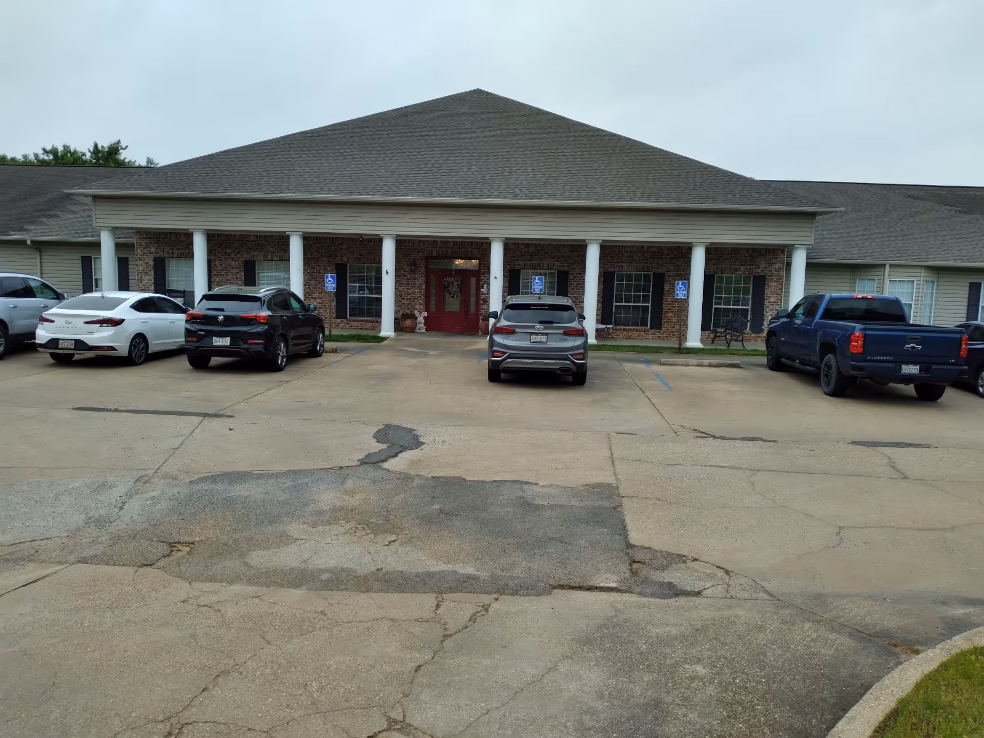 Front exterior view of a single-story assisted living community building with a covered entrance supported by white columns. Several cars are parked in front, including some in handicap parking spaces. The building has brick and siding exterior with multiple windows and a red door at the entrance.