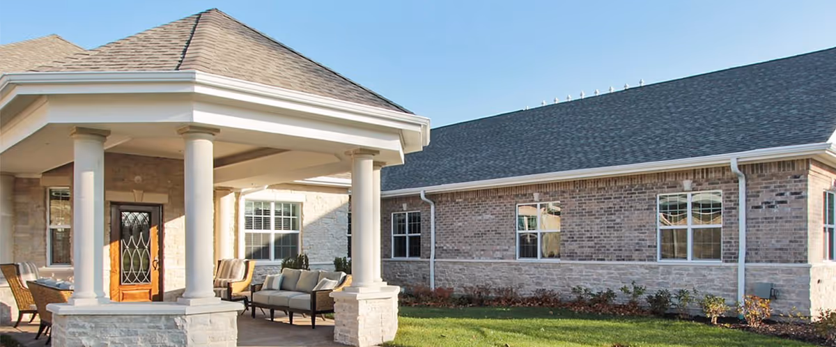 Front entrance of a brick memory care building with a covered portico, columns, outdoor seating and a lawn.