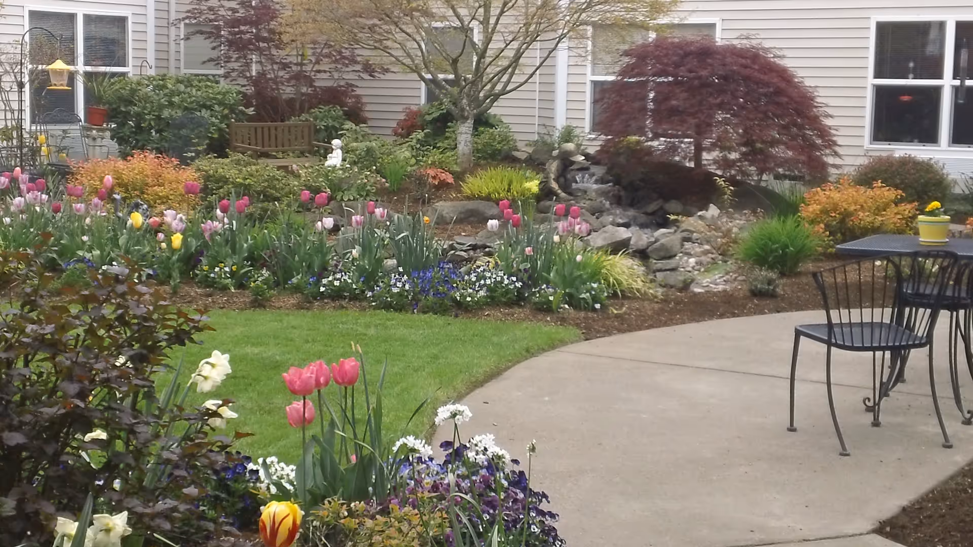 A landscaped courtyard with blooming tulips, a rock water feature, patio chairs and tables, and building windows in the background.