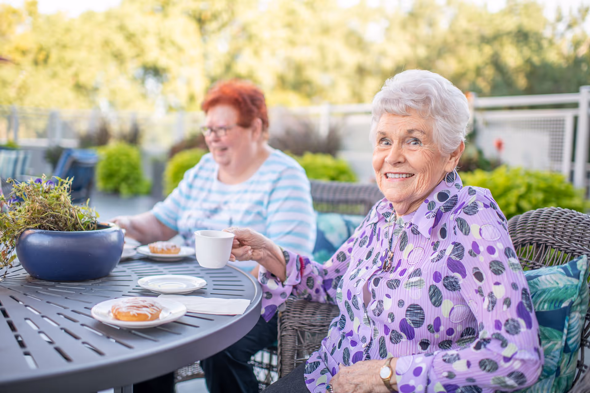 Two elderly women sitting outdoors at a round table with a potted plant, enjoying coffee and pastries. One woman in a purple patterned blouse is smiling at the camera, while the other woman in a striped shirt is seated in the background. The setting is a patio area with greenery and a railing in the background.