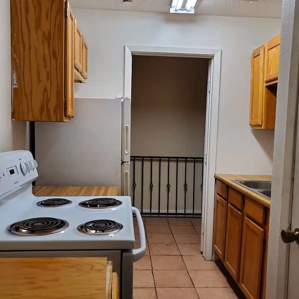 Narrow kitchen with an electric stove, wooden cabinets, a sink, and tile floor leading to a doorway with a metal railing.