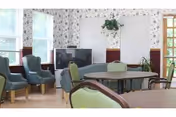 A common area in a senior living facility with several green and blue cushioned chairs arranged around tables. There is a television set against the wall, floral wallpaper, and large windows letting in natural light. A potted plant hangs on the wall above the television.