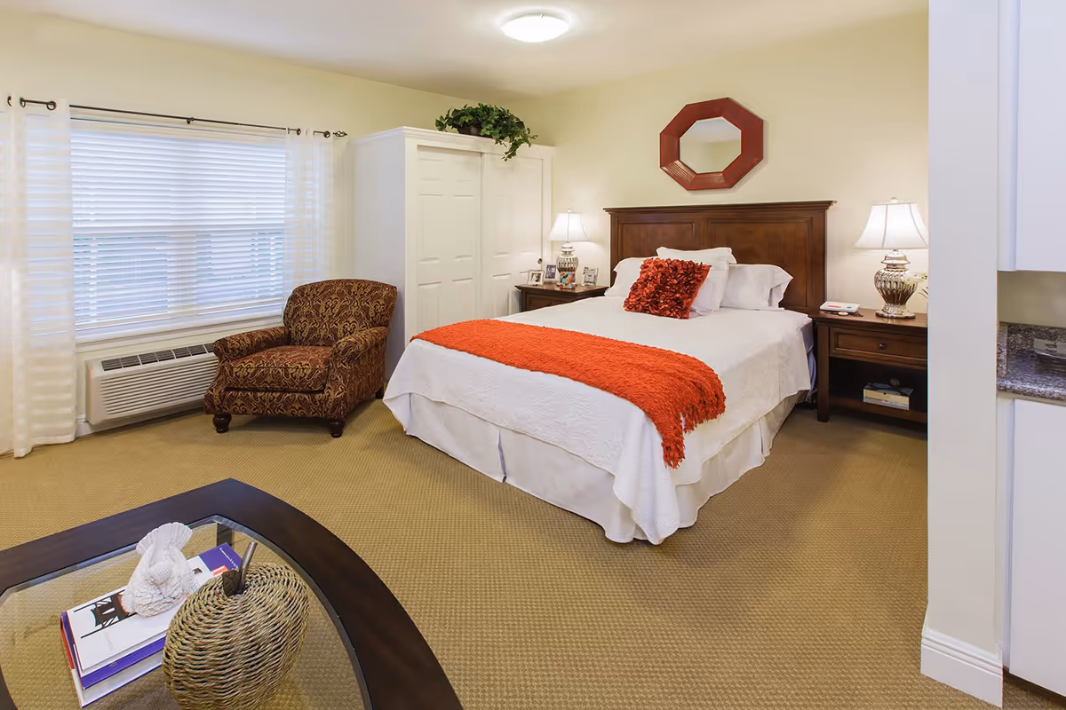 A cozy bedroom with a large bed dressed in white linens and a red throw blanket and pillow. There is a wooden headboard with a red octagonal mirror above it. Two bedside tables with lamps flank the bed. To the left, there is a patterned armchair near a window with white blinds and sheer curtains. A white wardrobe is positioned next to the chair. In the foreground, part of a glass-top coffee table with decorative items is visible.