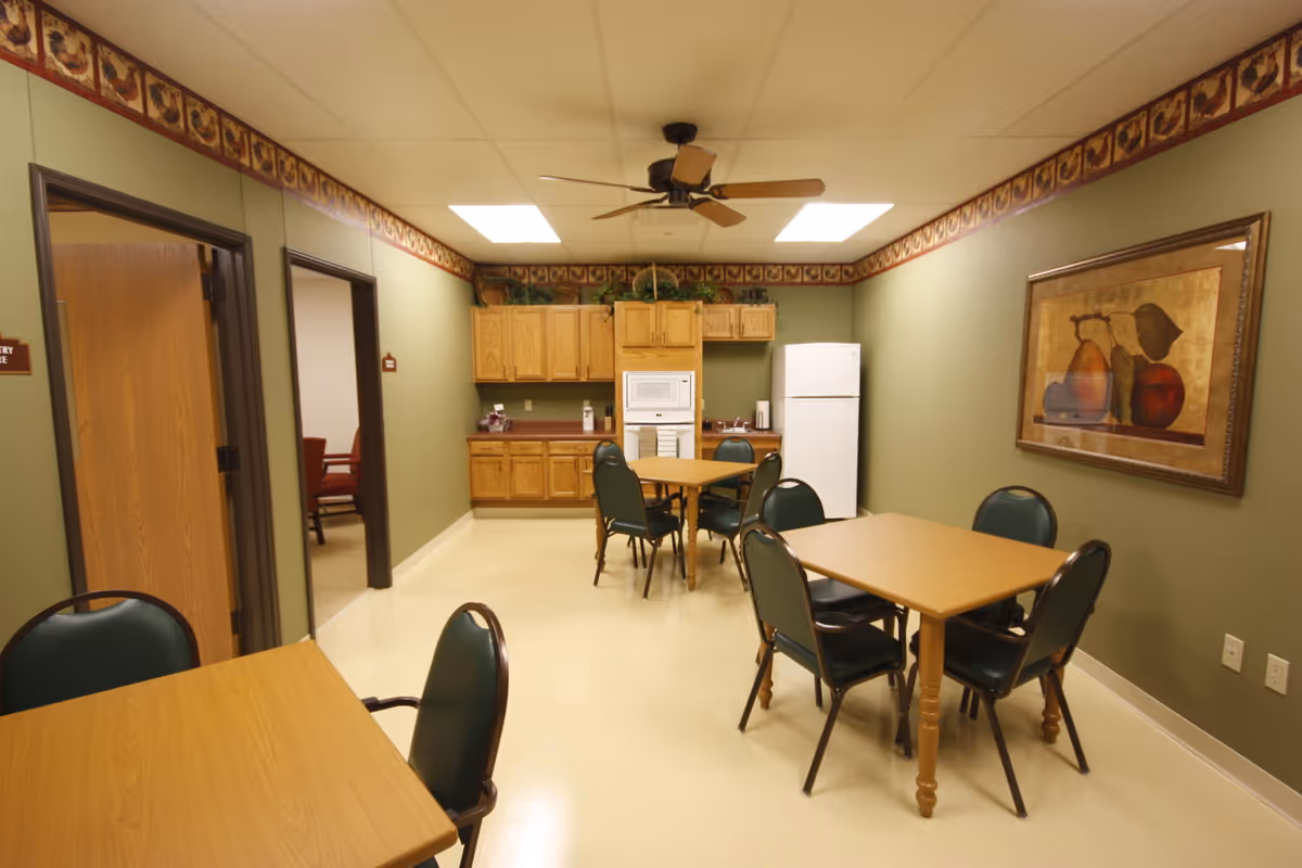 A small dining area with two wooden tables and green cushioned chairs. The room has light green walls with a decorative border near the ceiling. At the back, there is a kitchenette with wooden cabinets, a white refrigerator, a microwave, and a sink. A ceiling fan with wooden blades is mounted on the ceiling. A framed painting of fruit hangs on the right wall.