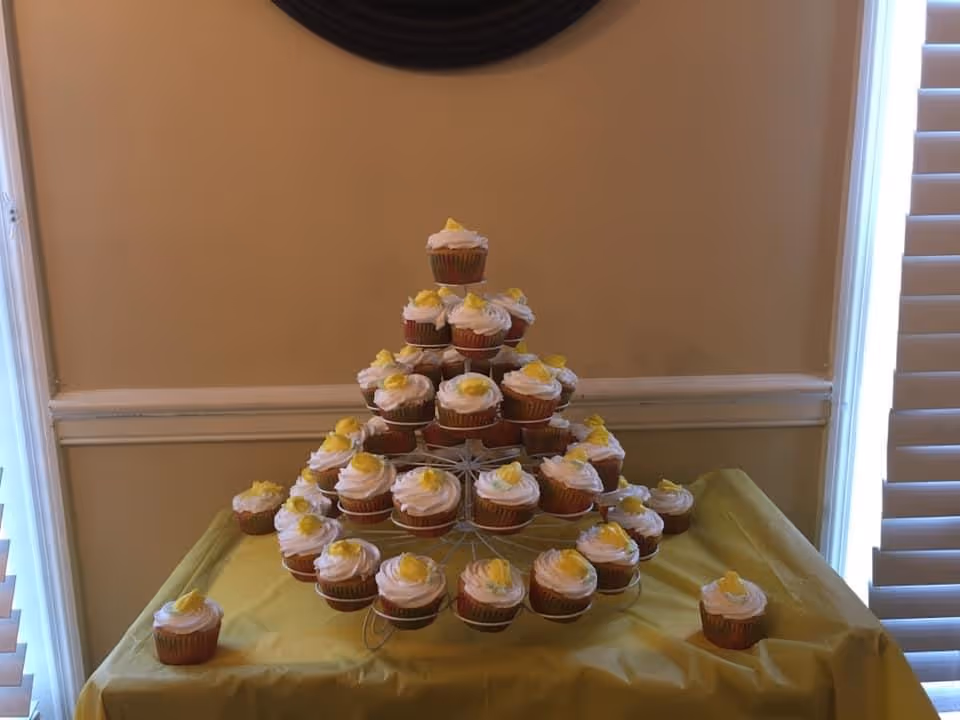 A tiered cupcake stand holding multiple cupcakes with white frosting and yellow decorations, placed on a yellow tablecloth-covered table in a room with beige walls and white window blinds.