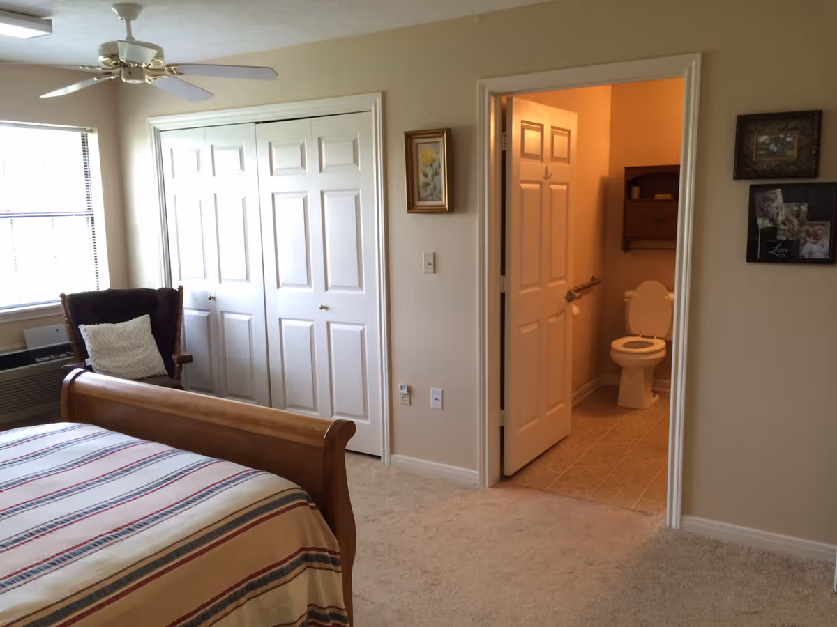 A bedroom with a wooden bed frame covered with a striped bedspread, a cushioned wooden chair with a white pillow, a ceiling fan, a window with blinds, and a closet with double white doors. An open door leads to a bathroom with a visible toilet and a wooden shelf above it. The walls are light-colored and there are framed pictures hanging on the walls.