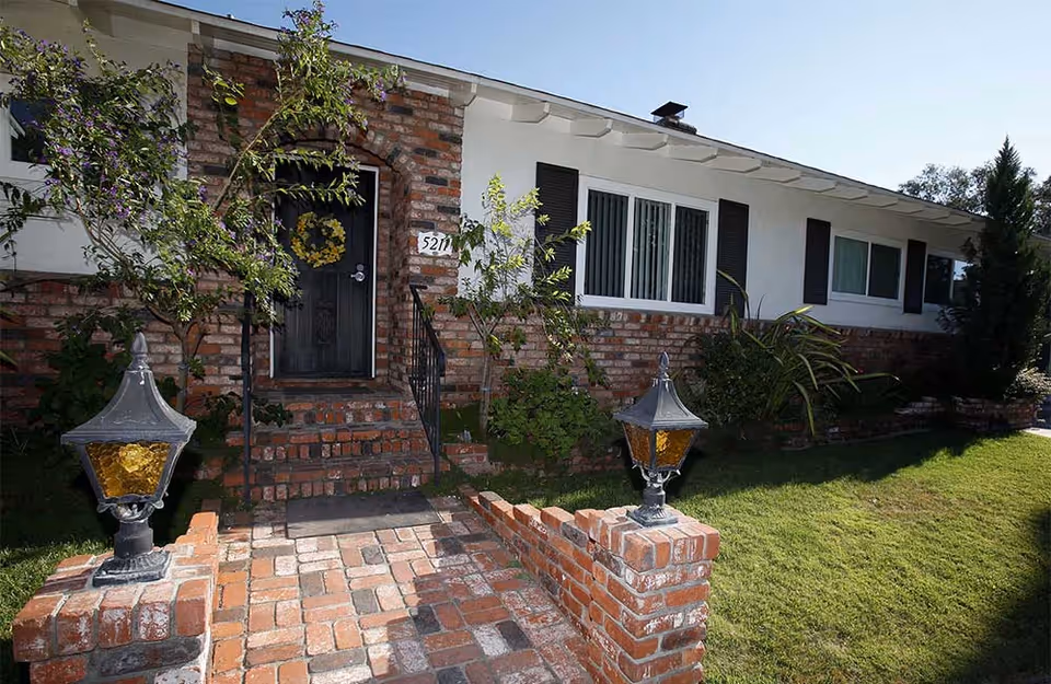 Front of a single-story brick house with a brick walkway, entrance steps, a black door with a wreath, and decorative lanterns.