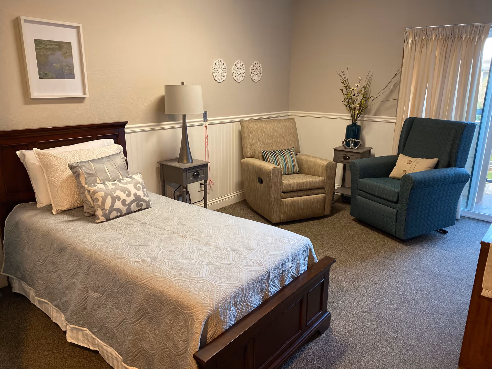 A cozy bedroom in a memory care facility featuring a single bed with multiple pillows and a light blue quilted bedspread. Next to the bed is a small nightstand with a modern lamp. The room also includes two comfortable armchairs, one beige and one blue, with decorative pillows. A small side table with a vase of flowers is positioned between the chairs. Light beige walls are decorated with framed artwork and three white decorative wall hangings. A sliding glass door with curtains allows natural light into the room.