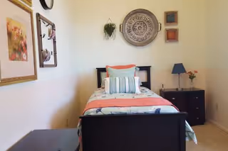 A small bedroom with a single bed against the wall, decorated with multiple pillows and a coral-colored blanket. The walls have framed artwork and decorative pieces, including a round ornamental piece and small square frames. There is a dark wooden nightstand with a lamp and a small vase with flowers beside the bed.