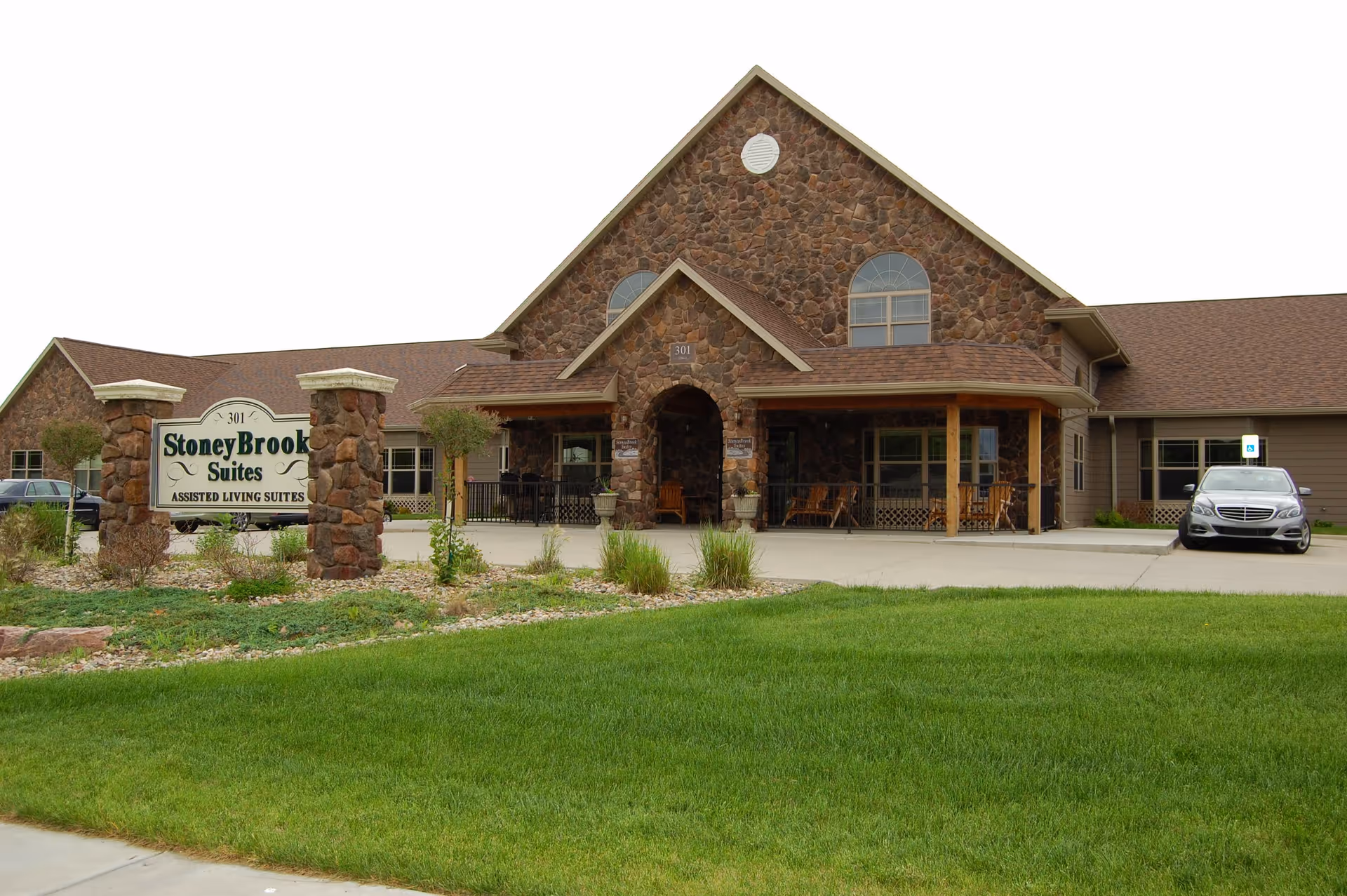 Exterior view of StoneyBrook Suites Assisted Living building with a stone facade, a covered entrance with wooden chairs, a green lawn in the foreground, and a sign displaying the facility name near the driveway where a silver car is parked.