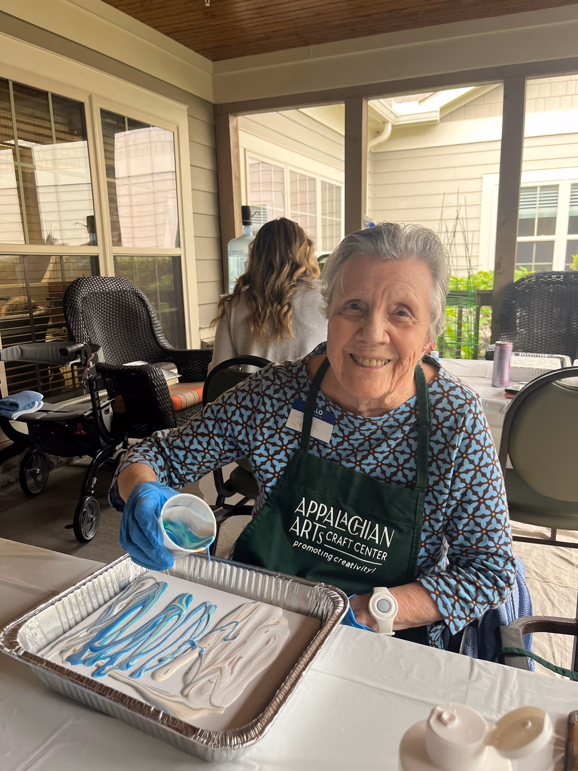 An elderly woman wearing a patterned shirt and a green Appalachian Arts Craft Center apron is smiling while pouring blue and white paint into a large aluminum tray. She is seated at a table covered with a white tablecloth, and there is a walker and another person with long hair sitting in the background on a covered porch area.