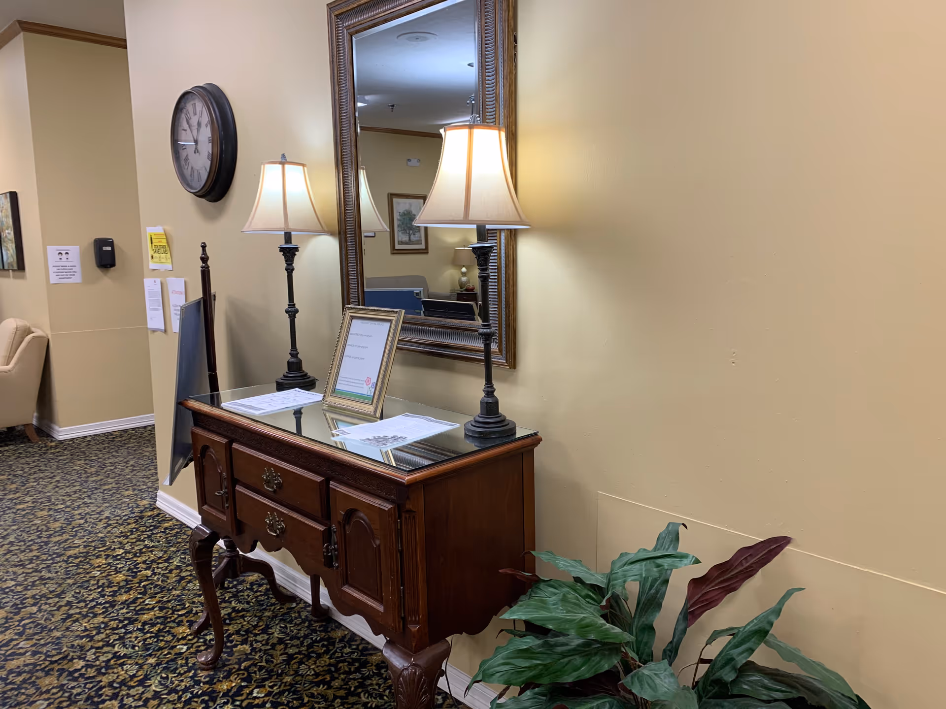 A hallway corner in a senior living facility featuring a wooden console table with two lamps and framed documents on top. Above the table is a large decorative mirror. A round wall clock is mounted on the adjacent wall, and a potted plant is placed on the floor next to the table. The walls are painted beige and the floor is covered with patterned carpet.
