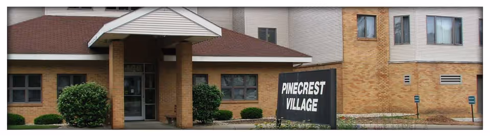 Exterior view of a brick building with a covered entrance supported by two brick pillars. There are bushes and small plants near the entrance. A black sign in front of the building reads 'PINECREST VILLAGE'.