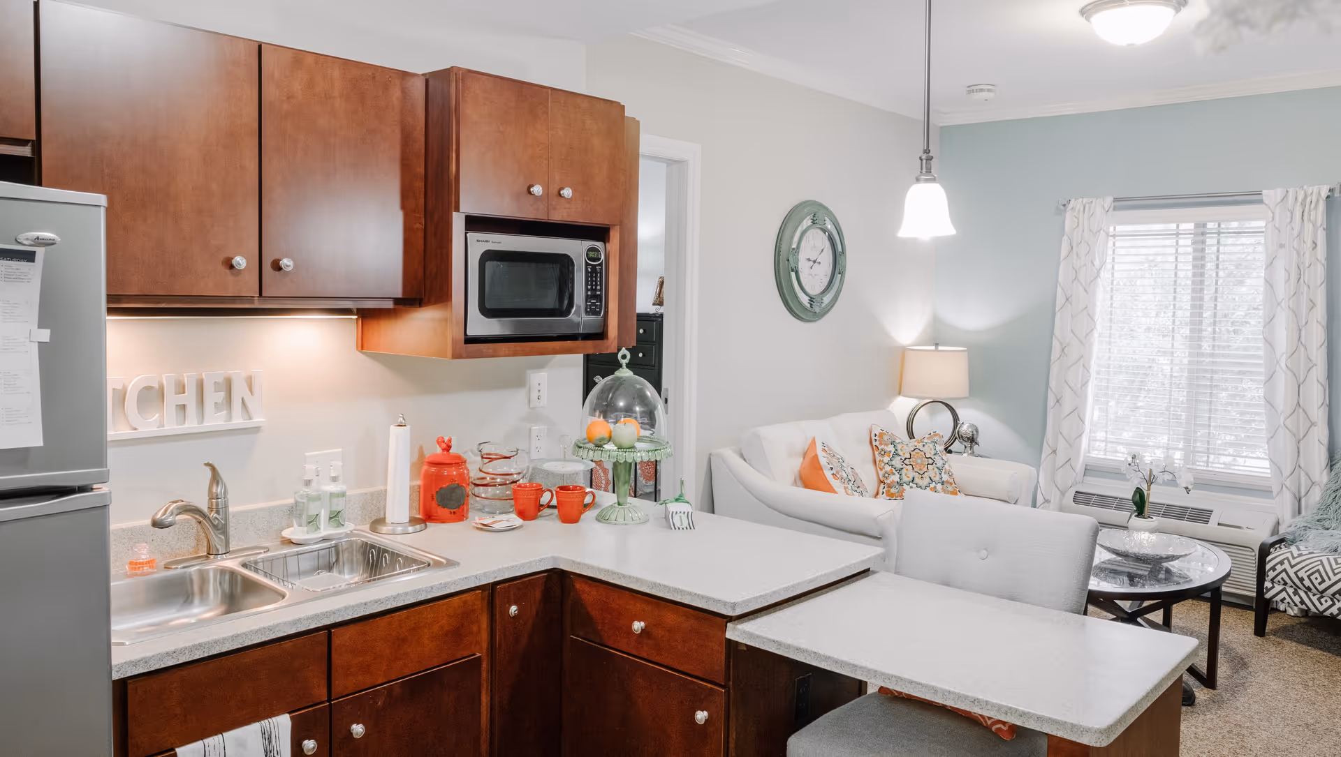 A cozy kitchen area with wooden cabinets, a stainless steel microwave, and a double sink. The countertop extends into a small dining area with a chair. In the background, there is a living room with a white couch, colorful pillows, a round glass coffee table, a window with white curtains, and a wall clock.