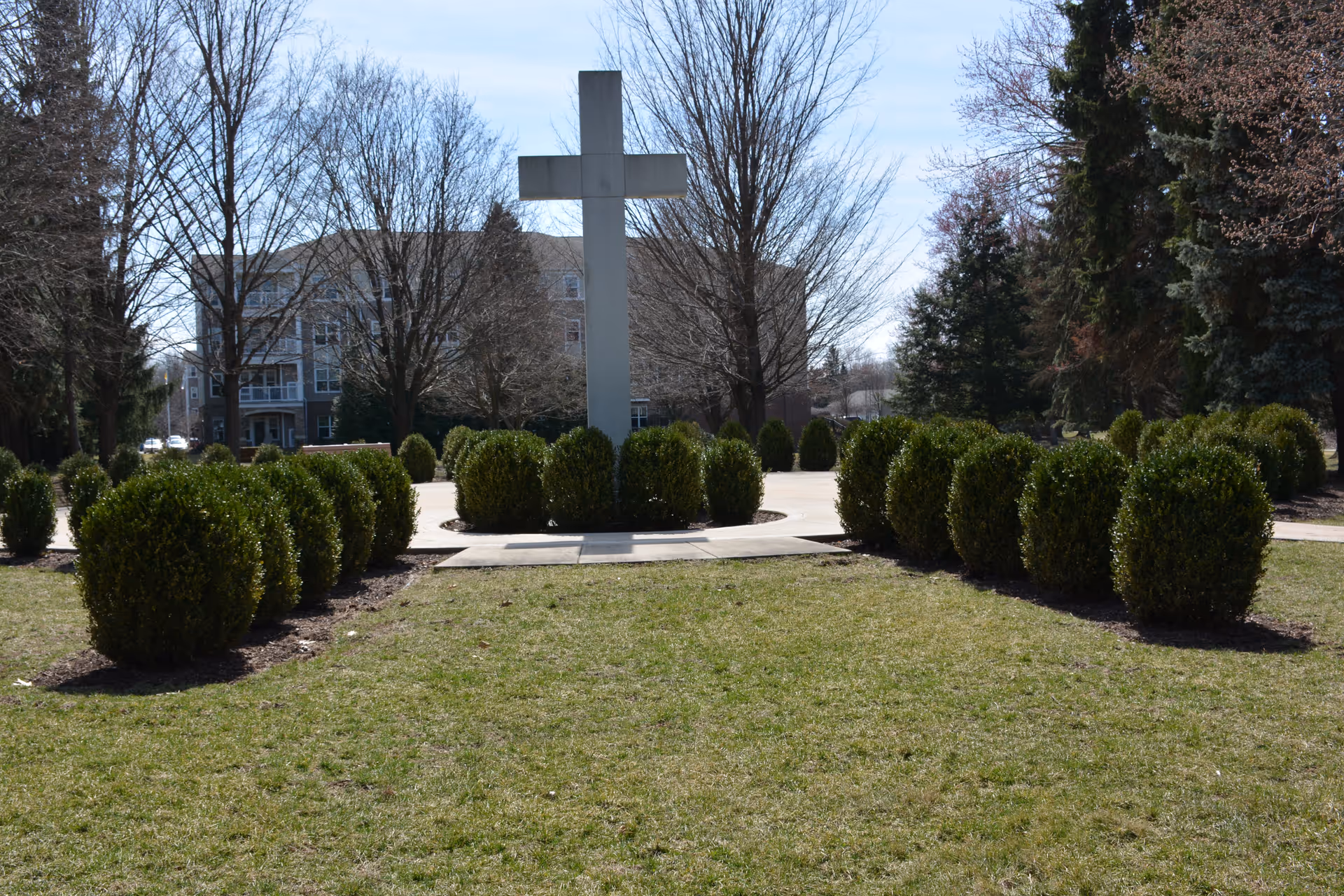 Outdoor view of a large stone cross surrounded by neatly trimmed bushes and trees with a multi-story building in the background under a partly cloudy sky.