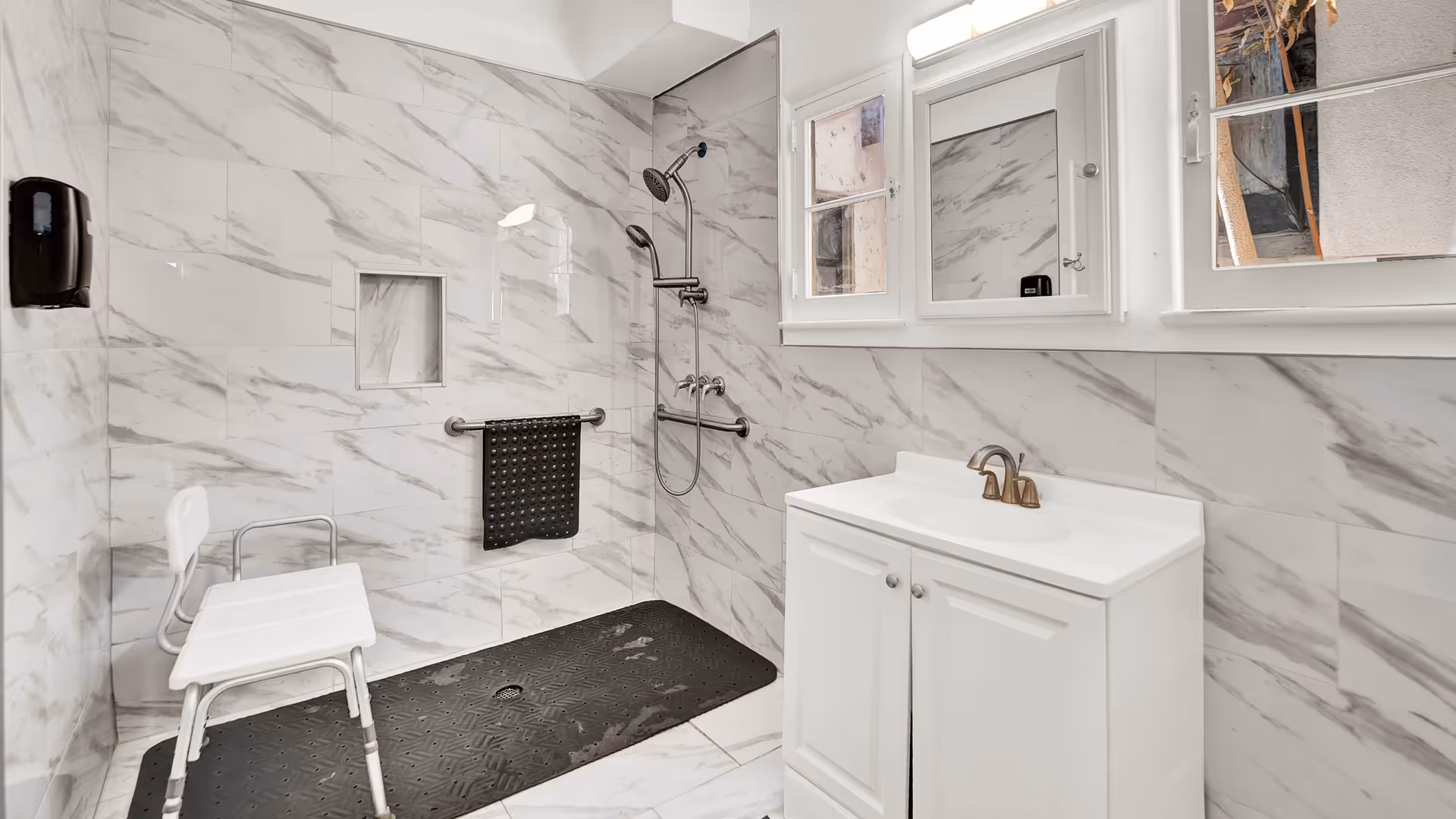 A modern bathroom with white and gray marble tiles covering the walls and floor. The shower area features a handheld showerhead and a fixed showerhead, with a black non-slip mat and a white shower chair. There is a white vanity with a sink and a mirror above it, and two windows letting in natural light.