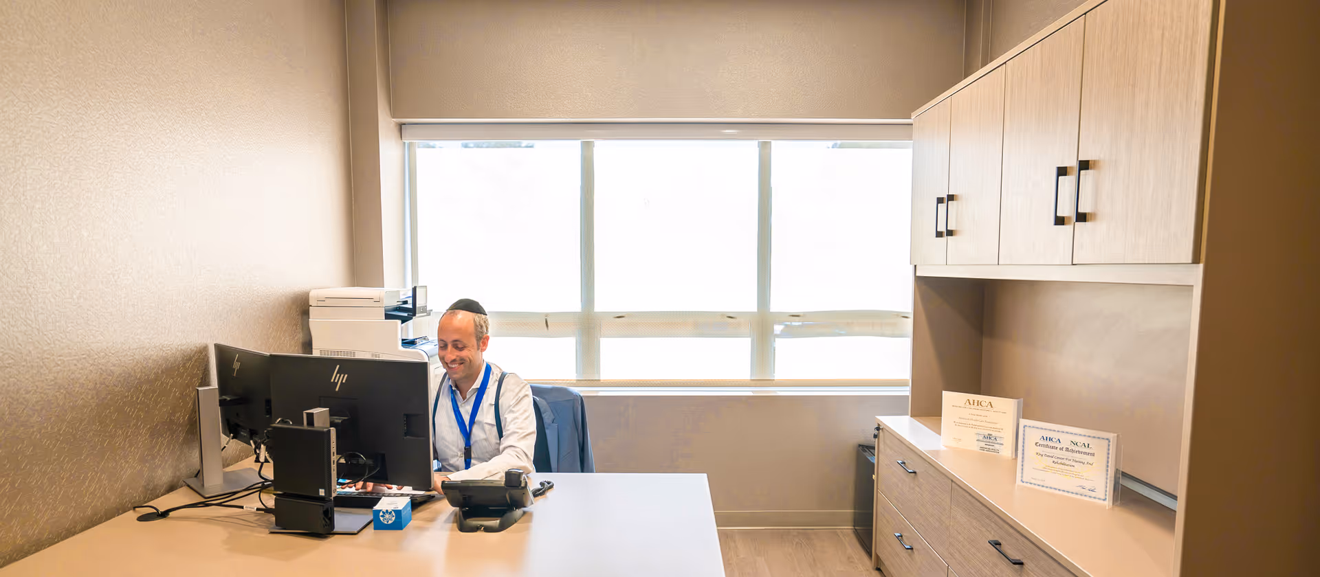 A man wearing a white shirt and blue lanyard is sitting at a desk in a well-lit office with a large window behind him. The desk has two computer monitors, a telephone, and office supplies. There are cabinets on the right side of the room with certificates displayed on the countertop.