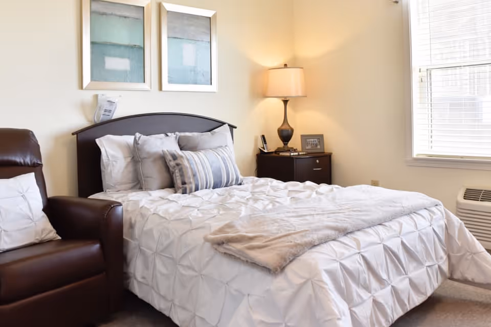 A bright, neatly made bedroom with a bed topped with decorative pillows, a bedside table and lamp, and a leather recliner by the window.