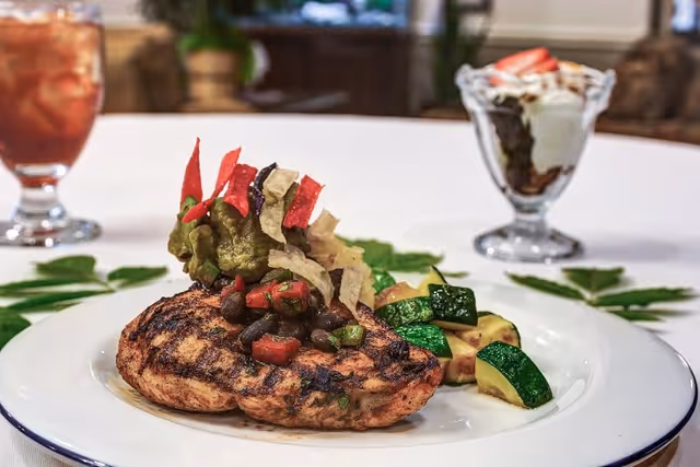 A plated meal featuring a grilled chicken breast topped with guacamole, black beans, diced tomatoes, and crispy tortilla strips, served with sautéed zucchini on the side. In the background, there is a glass of iced tea and a dessert parfait with whipped cream and strawberries on a white tablecloth.