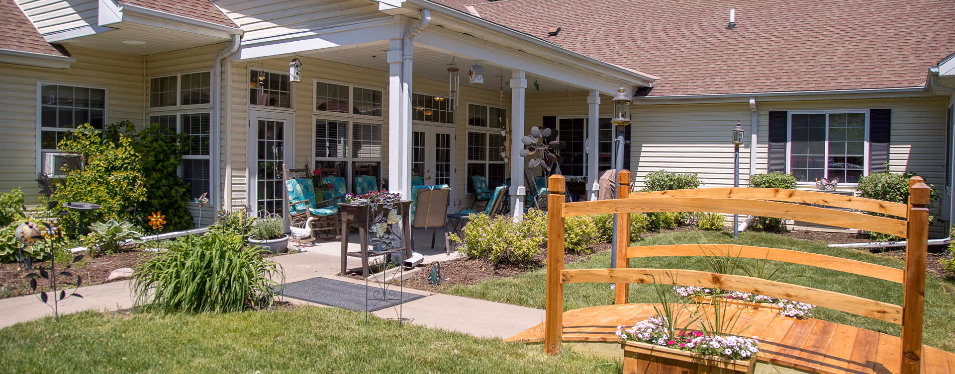Sunlit courtyard with a wooden garden bridge and patio seating in front of a single-story senior living building.
