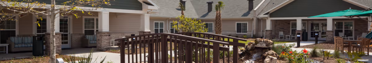 Exterior courtyard of a senior living facility featuring a wooden bridge, seating areas, landscaping, and a covered patio.