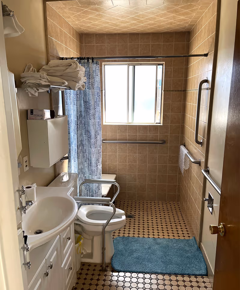A bathroom with beige tiled walls and floor, featuring a white sink with cabinet, a toilet with safety rails, a blue bath mat, a shower area with a blue and white patterned curtain, grab bars on the walls, and a window letting in natural light.
