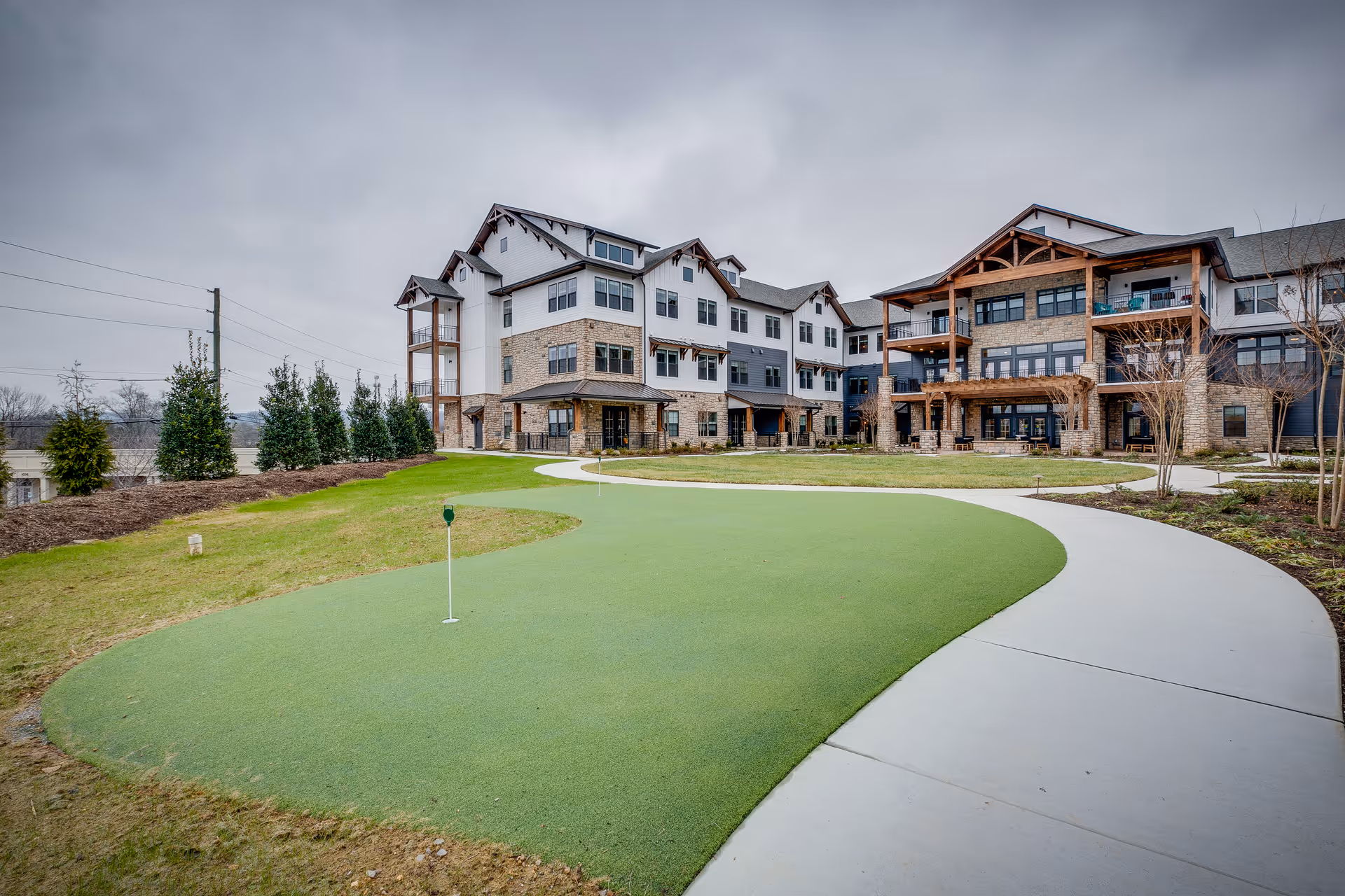 Exterior view of a multi-story senior living facility with a putting green and a curved concrete walkway in the foreground. The building features a combination of stone and siding with balconies and large windows under a cloudy sky.