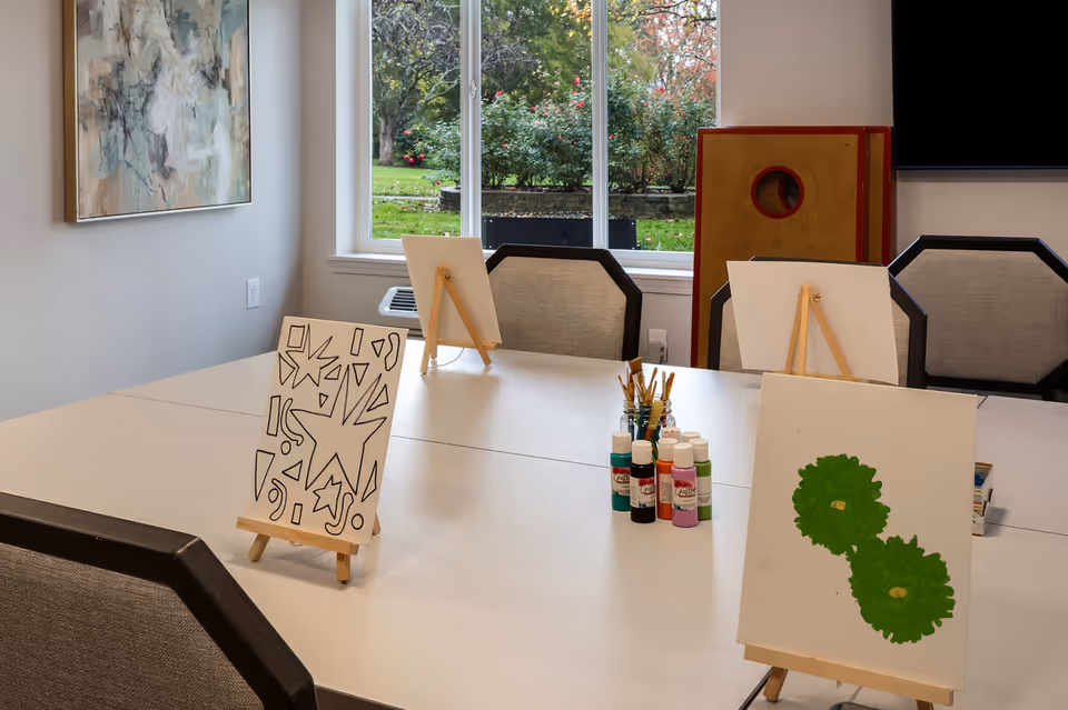 A bright room with a large white table set up for painting activities. There are three small easels on the table, each holding a canvas with different artwork: one with black abstract shapes, one with two green circular shapes, and one blank. Several bottles of paint and jars of paintbrushes are arranged in the center of the table. The room has a window showing a green garden outside, a piece of abstract wall art, and several cushioned chairs around the table.