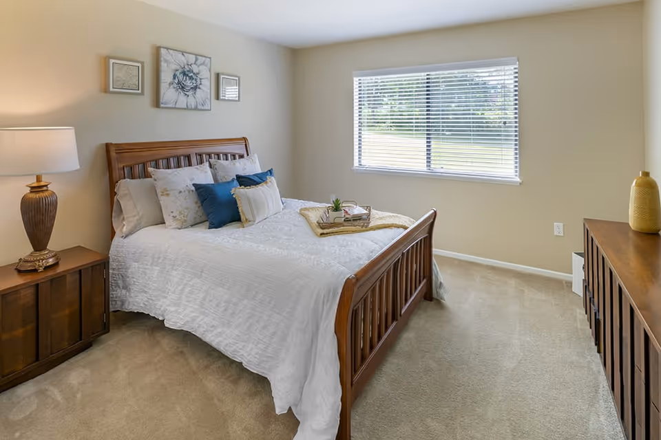A neatly made bed with white bedding and multiple pillows in a bedroom. There is a wooden nightstand with a lamp on the left side of the bed and a wooden dresser on the right side of the room. A window with blinds lets in natural light, and three pieces of wall art hang above the bed.