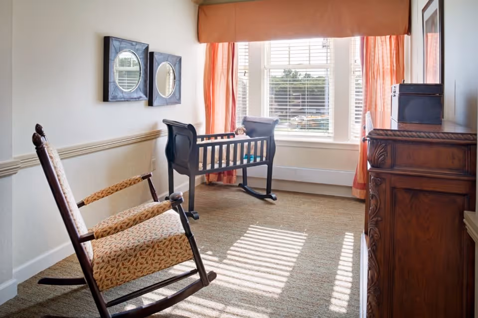 A cozy room with a wooden rocking chair, a dark wooden cradle with a doll inside, two framed mirrors on the wall, and a wooden dresser. The room has a large window with white blinds and orange curtains, allowing sunlight to cast shadows on the carpeted floor.