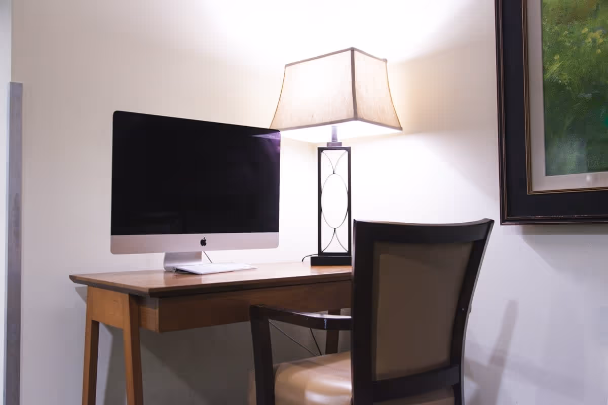 A wooden desk with a large Apple iMac computer and a modern table lamp with a rectangular beige shade. A cushioned chair with wooden frame is positioned in front of the desk. A framed painting with green tones hangs on the white wall to the right.