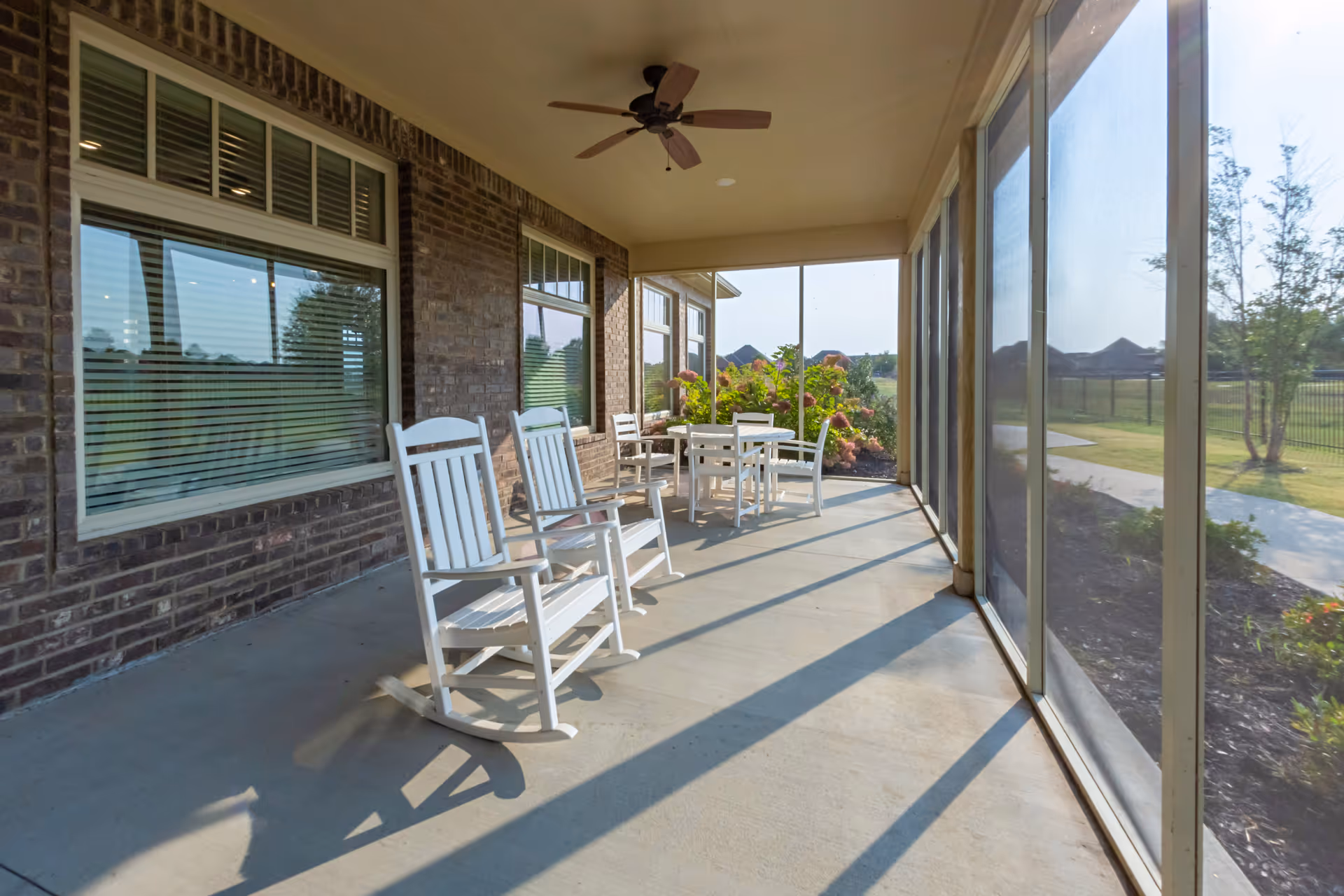 A screened-in porch area with two white rocking chairs and a white table with four chairs. The porch has a ceiling fan and overlooks a garden with flowers and a grassy area beyond a fence.